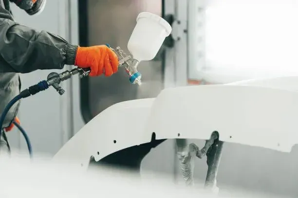 Person sprays white paint on a car bumper with a spray gun, wearing protective gear.
