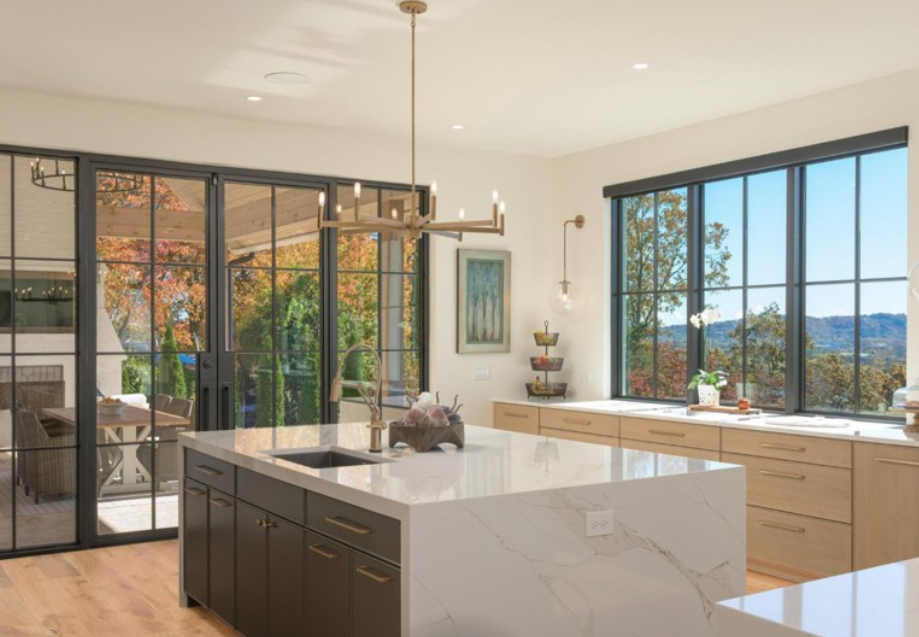 Modern kitchen with large island, black-framed windows, and a view of trees and sky.