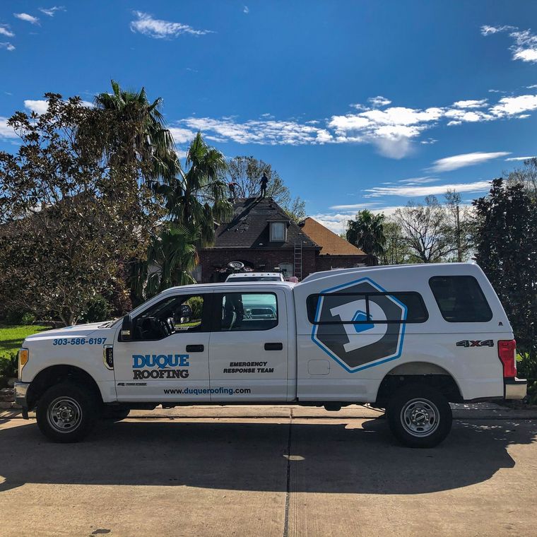 White Duque Roofing truck parked, house with ladder, blue sky.