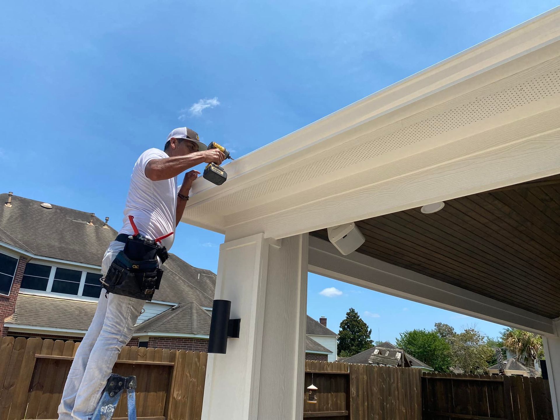 Man on ladder installing a light-colored fascia on a patio roof with a drill. Bright sunny day.