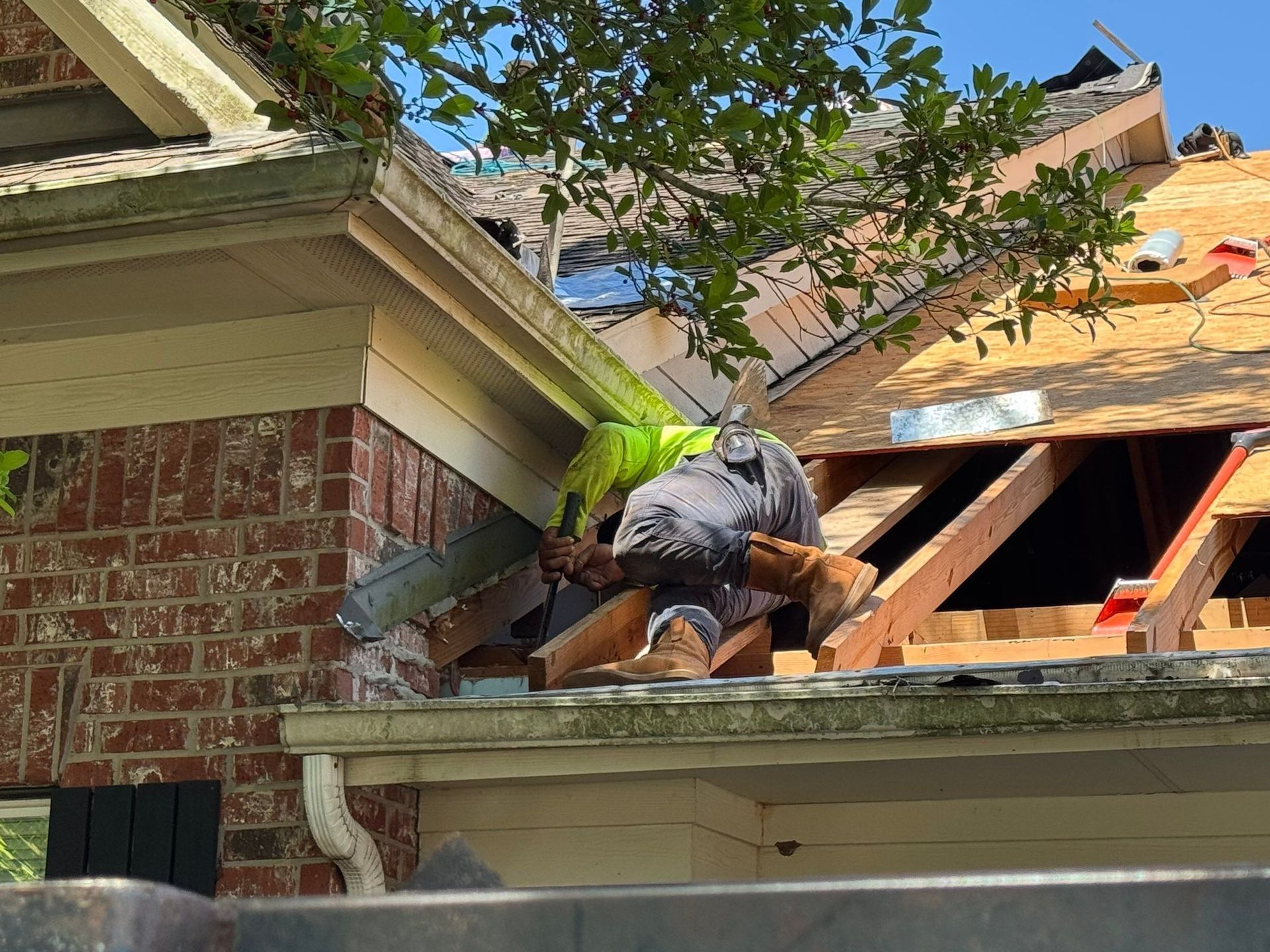 Construction worker on a roof, removing shingles. Exposed rafters and brick siding are visible.