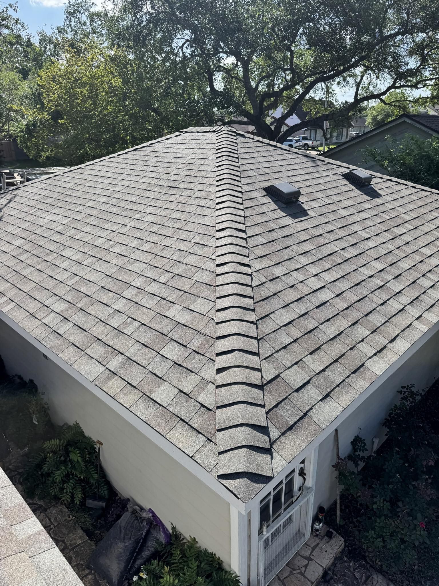 Overhead view of a roof with weathered gray shingles, a central ridge, and two small vents on a sunny day.