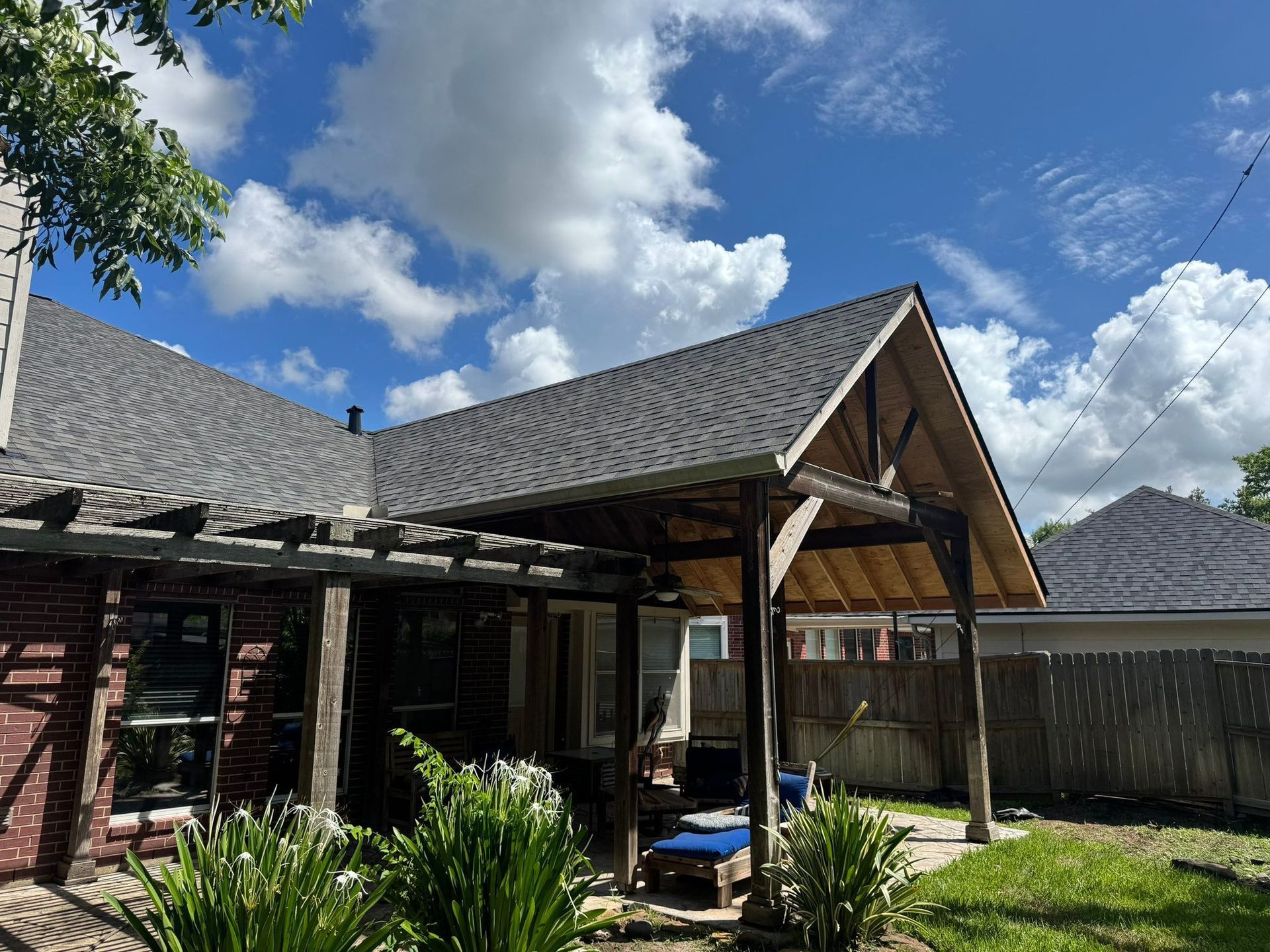 Backyard with covered patio and brick house, under a cloudy blue sky.