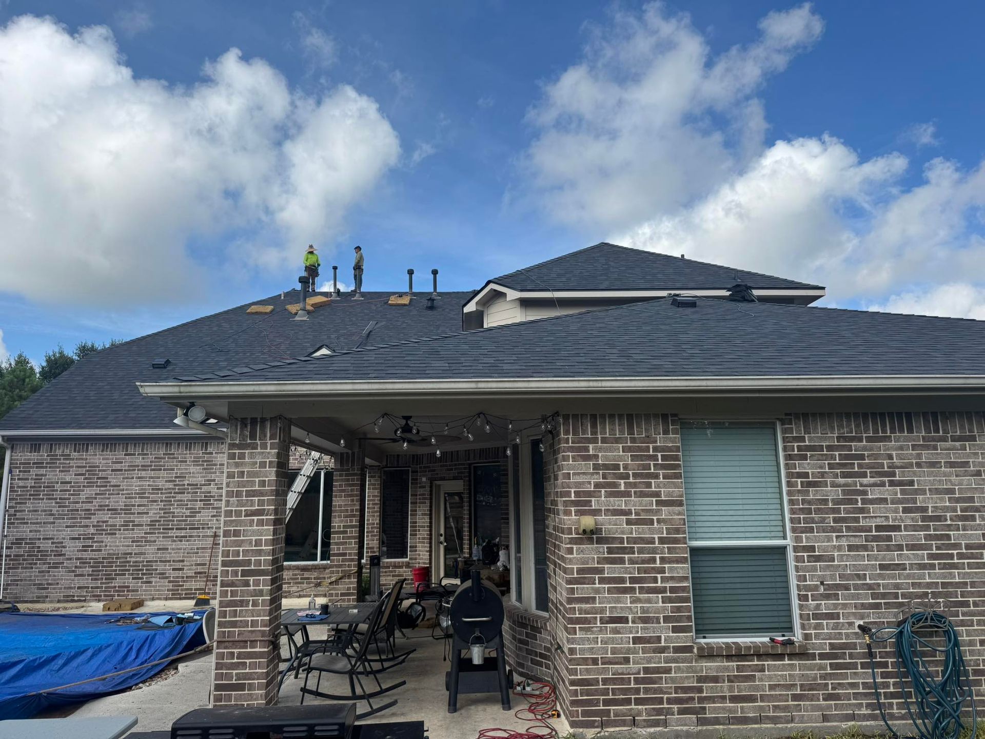 Workers on a dark-shingled roof under a partly cloudy sky; brick house with a covered patio and pool.