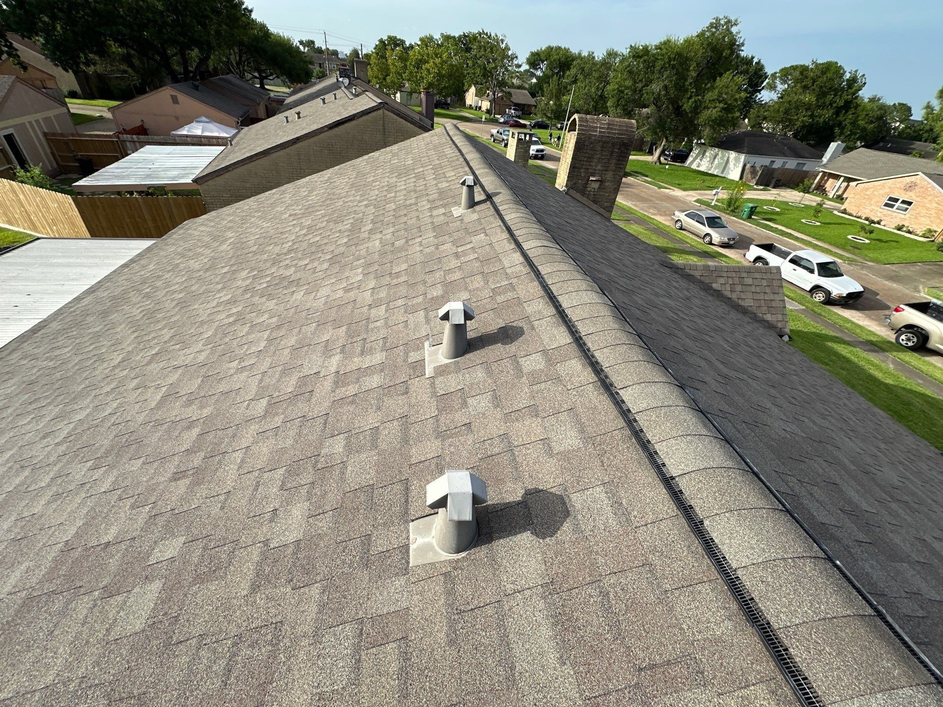 View from a rooftop. Brown shingle roof with vents, overlooking a residential neighborhood with houses and cars.
