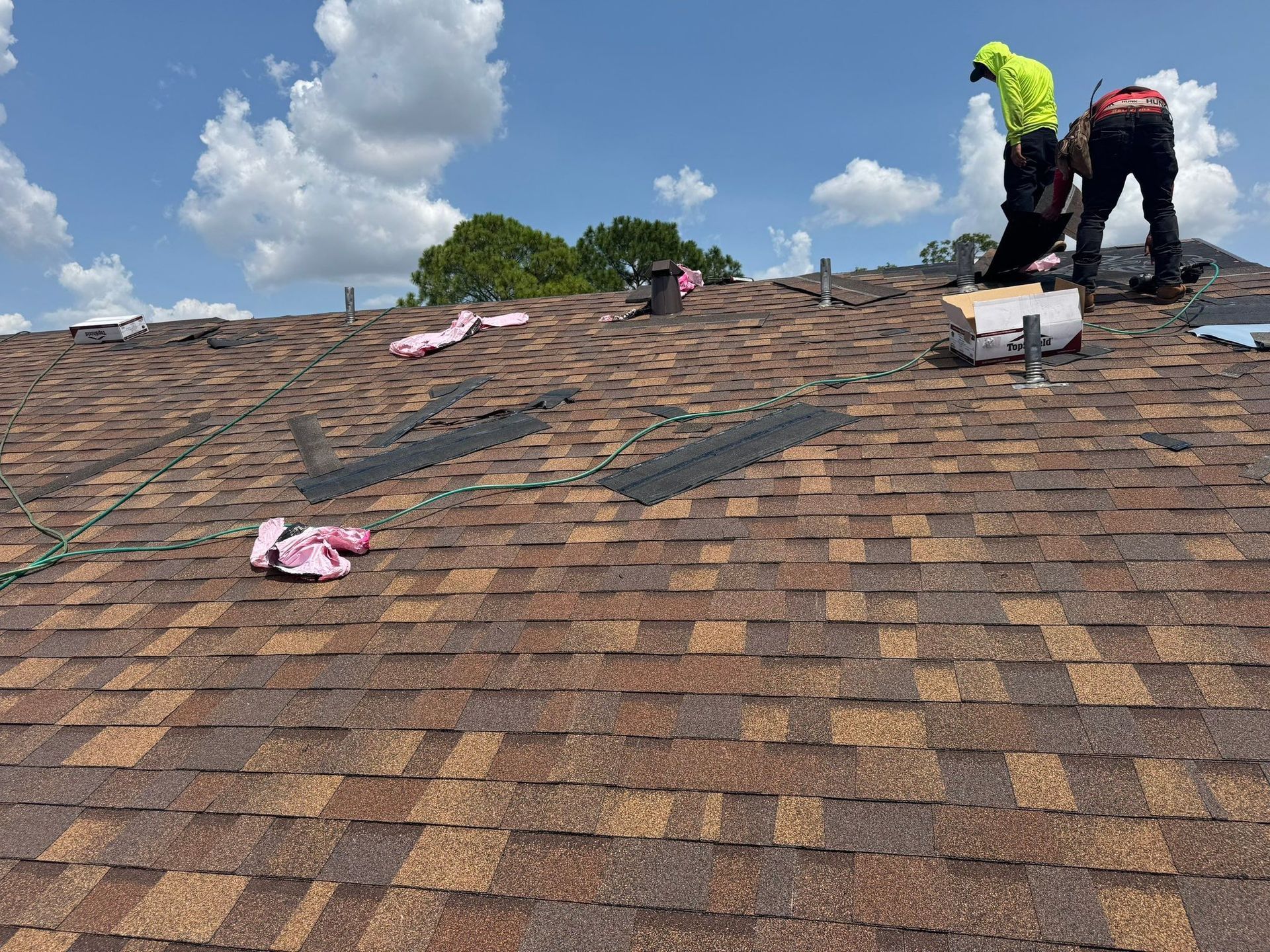 Two workers on a brown shingle roof under a blue sky, repairing sections with new shingles.