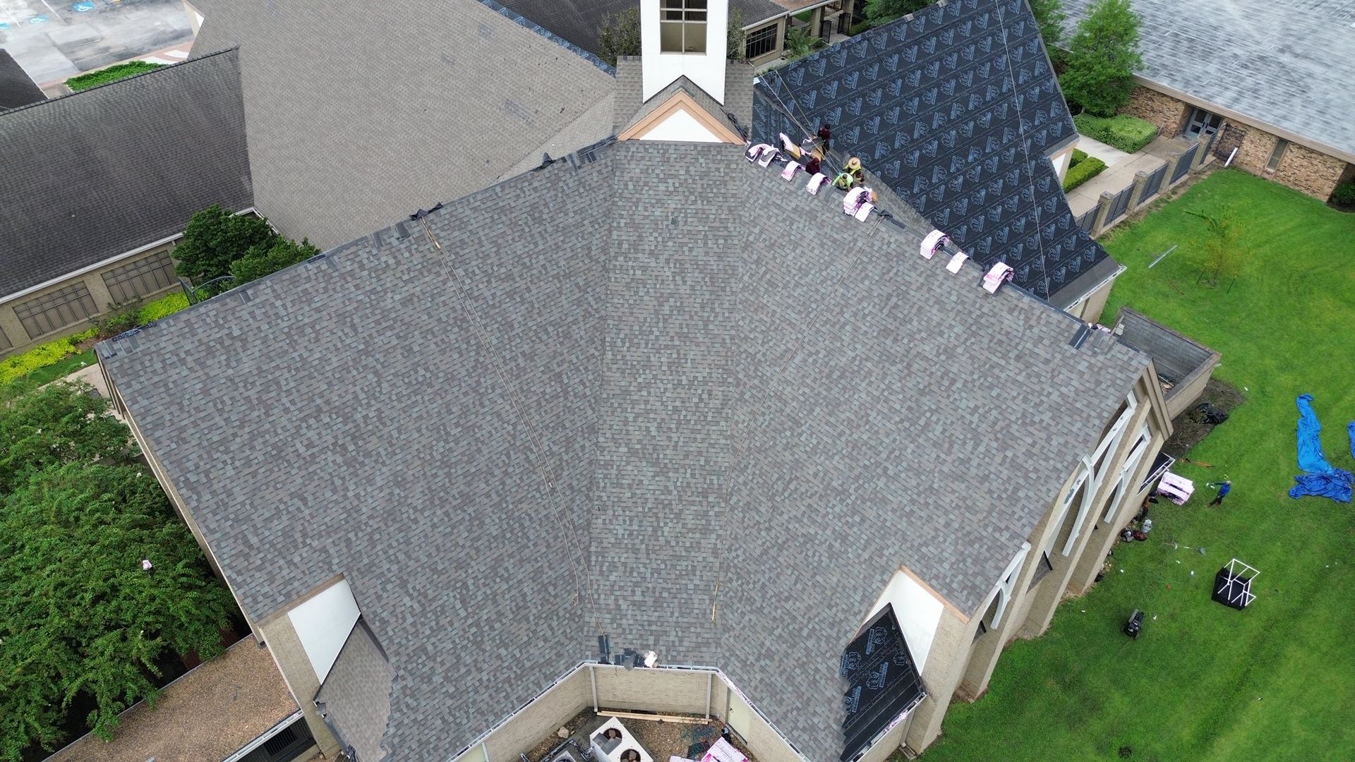 Aerial view of workers on a shingled roof, some removing old shingles, some installing new ones.