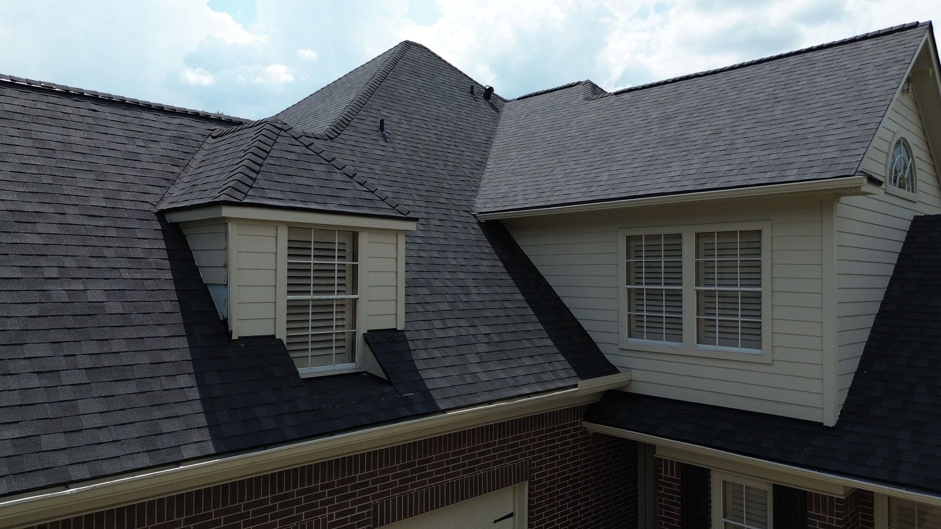 Roof of a house with dark gray shingles, dormer windows, and a brick facade.