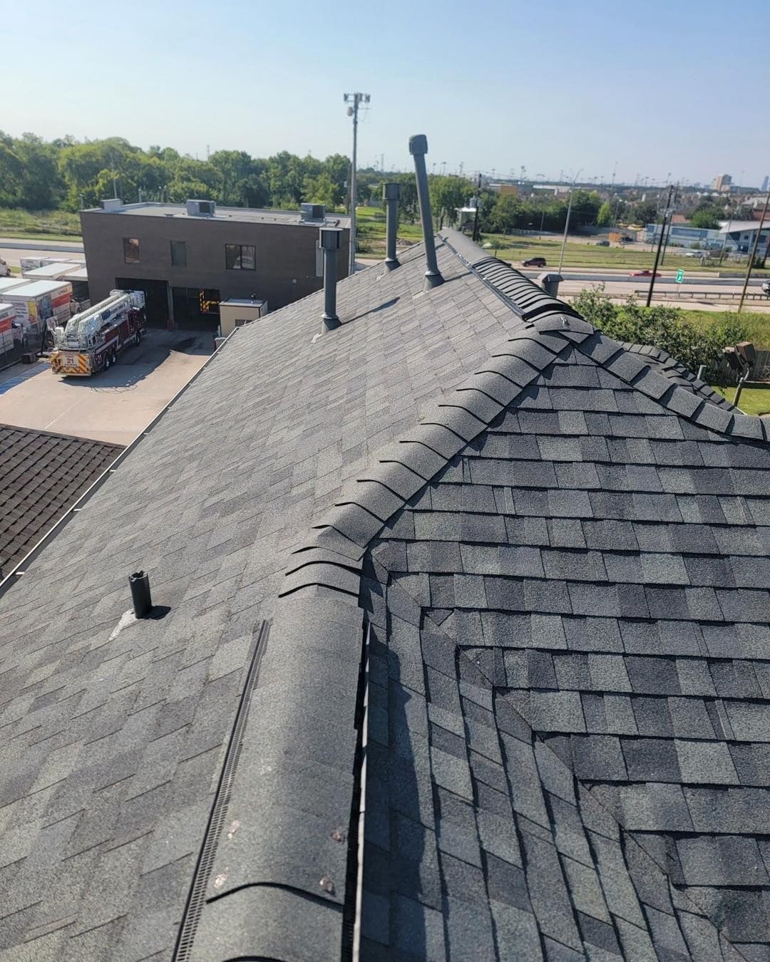 Gray asphalt shingle roof with chimneys and a commercial building in the background on a sunny day.