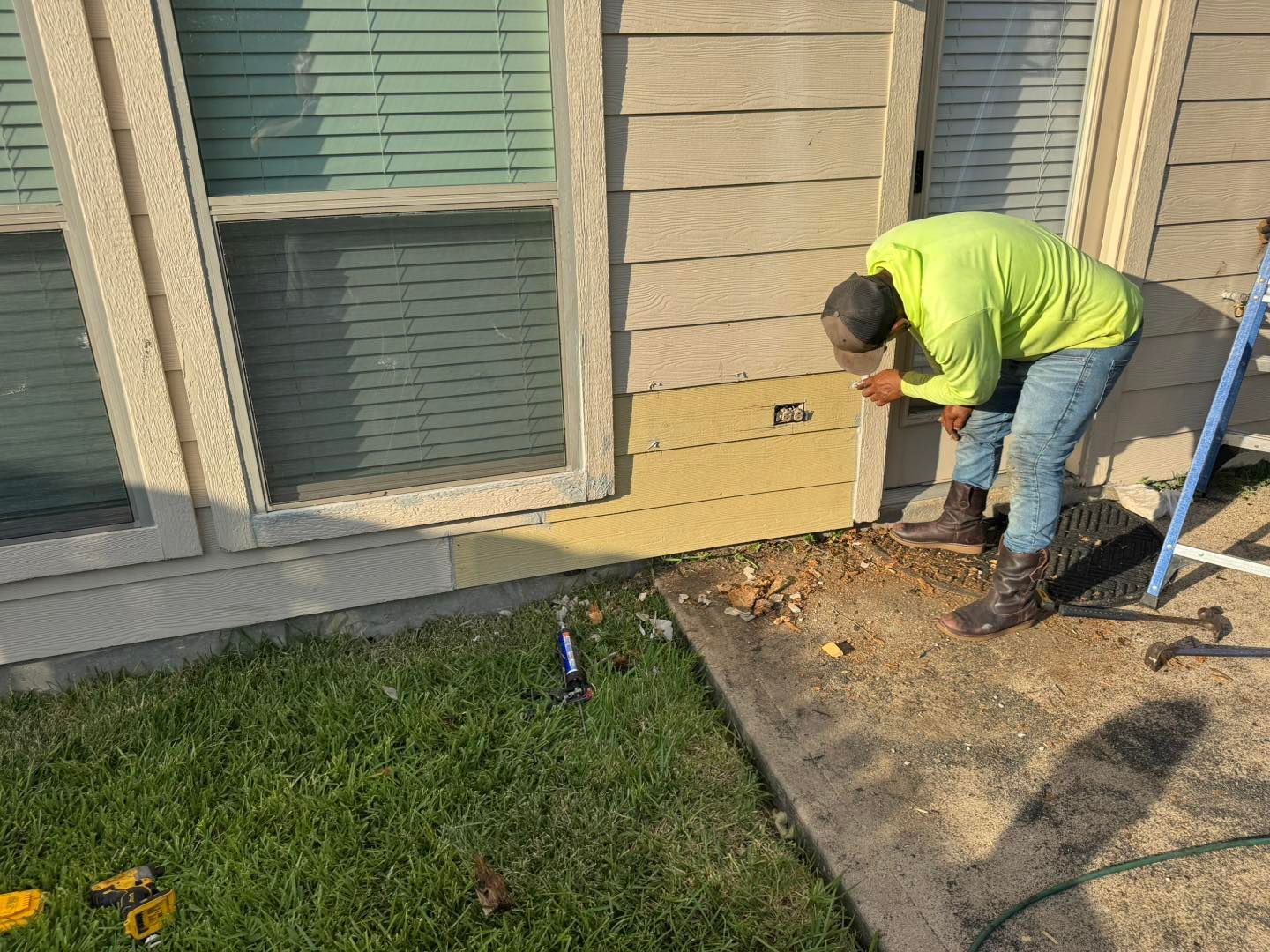 Person in neon green shirt working on siding near a window and concrete patio.
