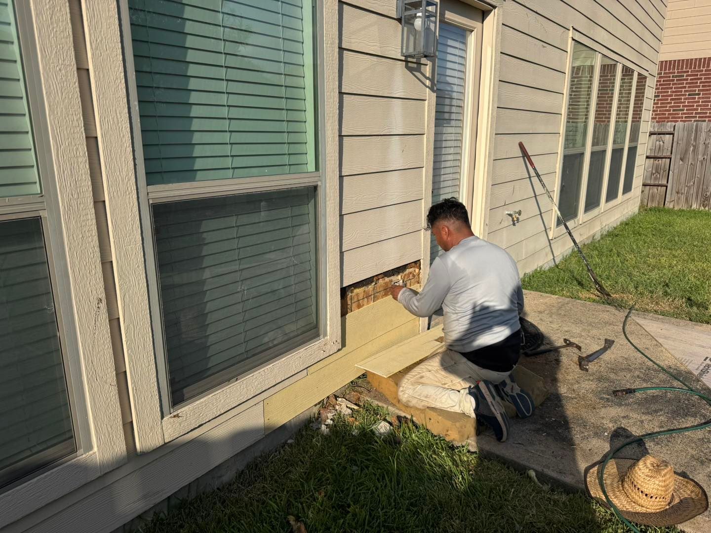 Person kneels, repairing siding near a window and door on a house. Beige siding, green grass, concrete path.
