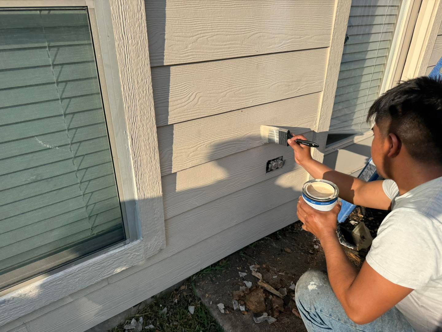 Person painting light-colored siding on a house exterior with a brush and paint can.