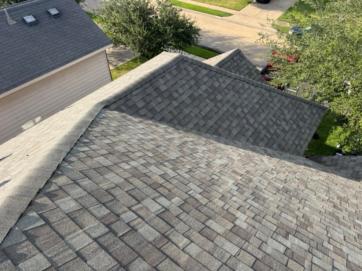 Overhead view of a roof covered in gray asphalt shingles, with a glimpse of a tan building and green trees.