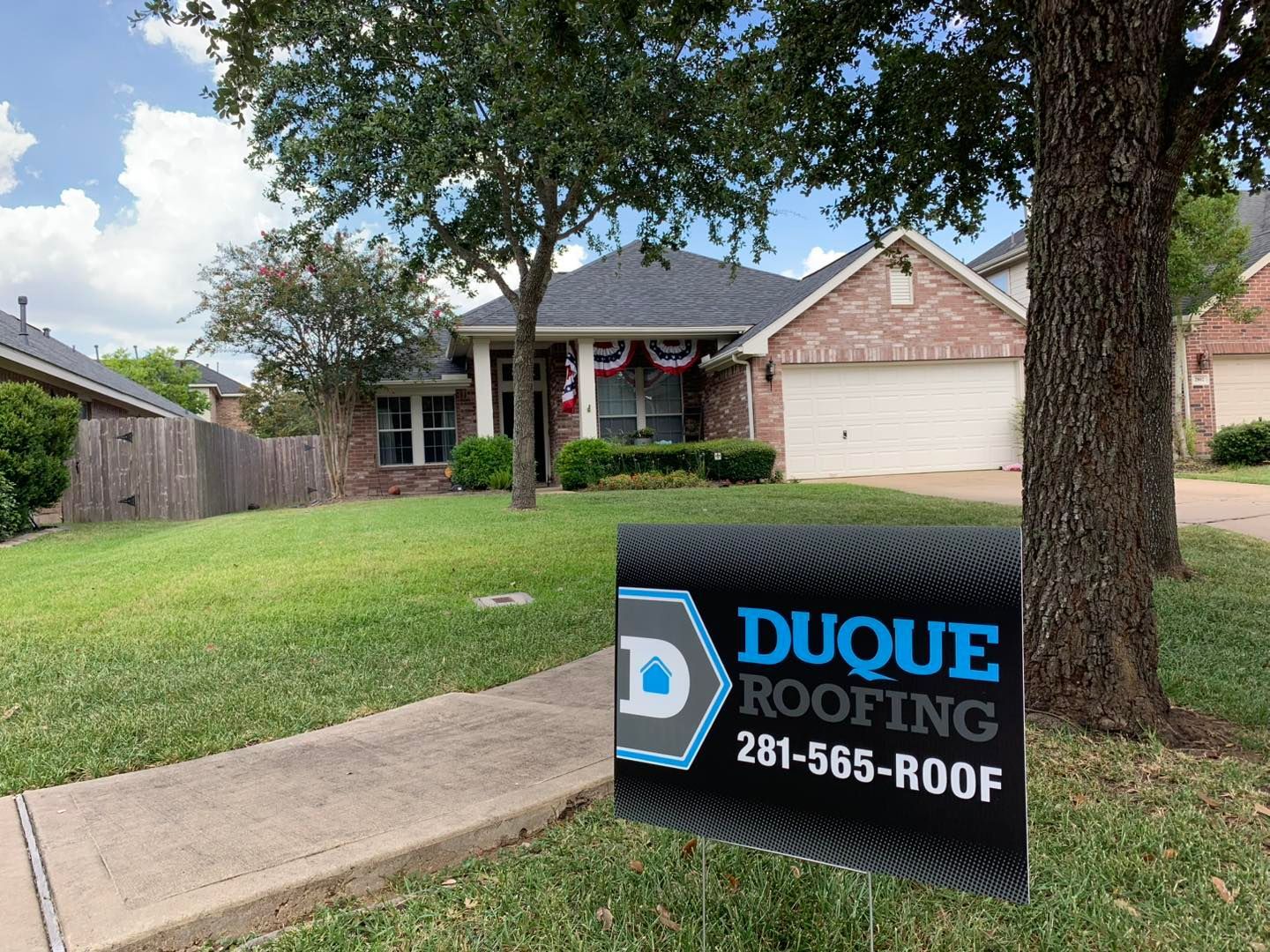 A house with a Duque Roofing sign on the lawn. A tree is in the foreground.
