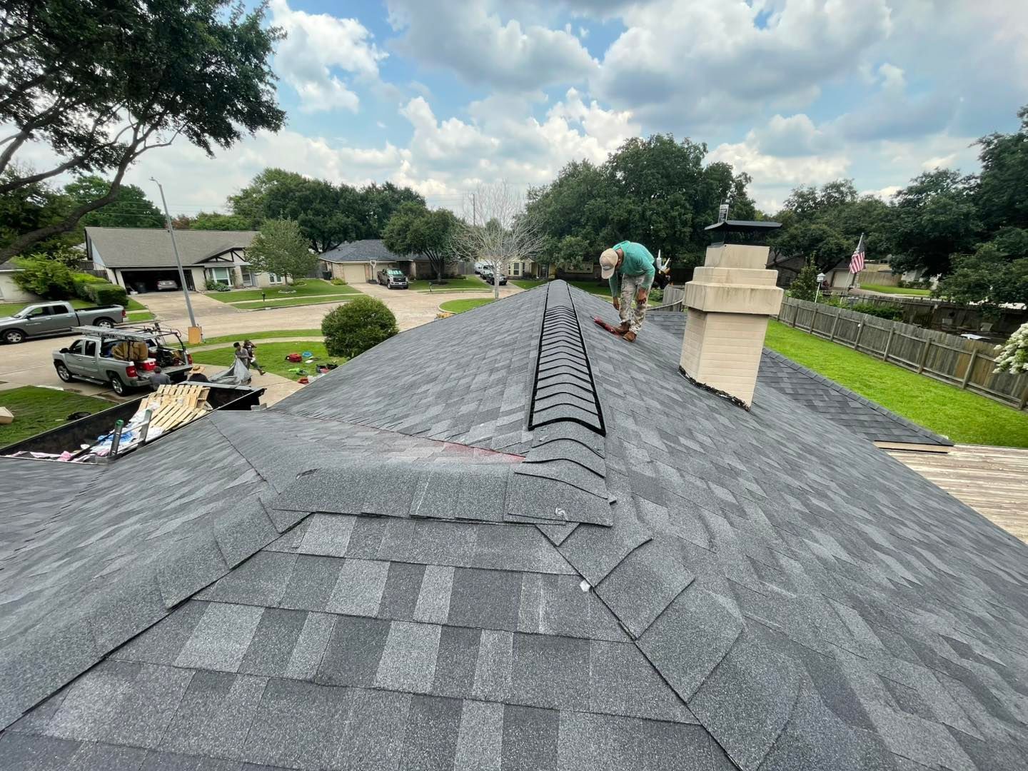 Roofer on a gray shingle roof under a cloudy sky. A chimney and roof ladder are present.