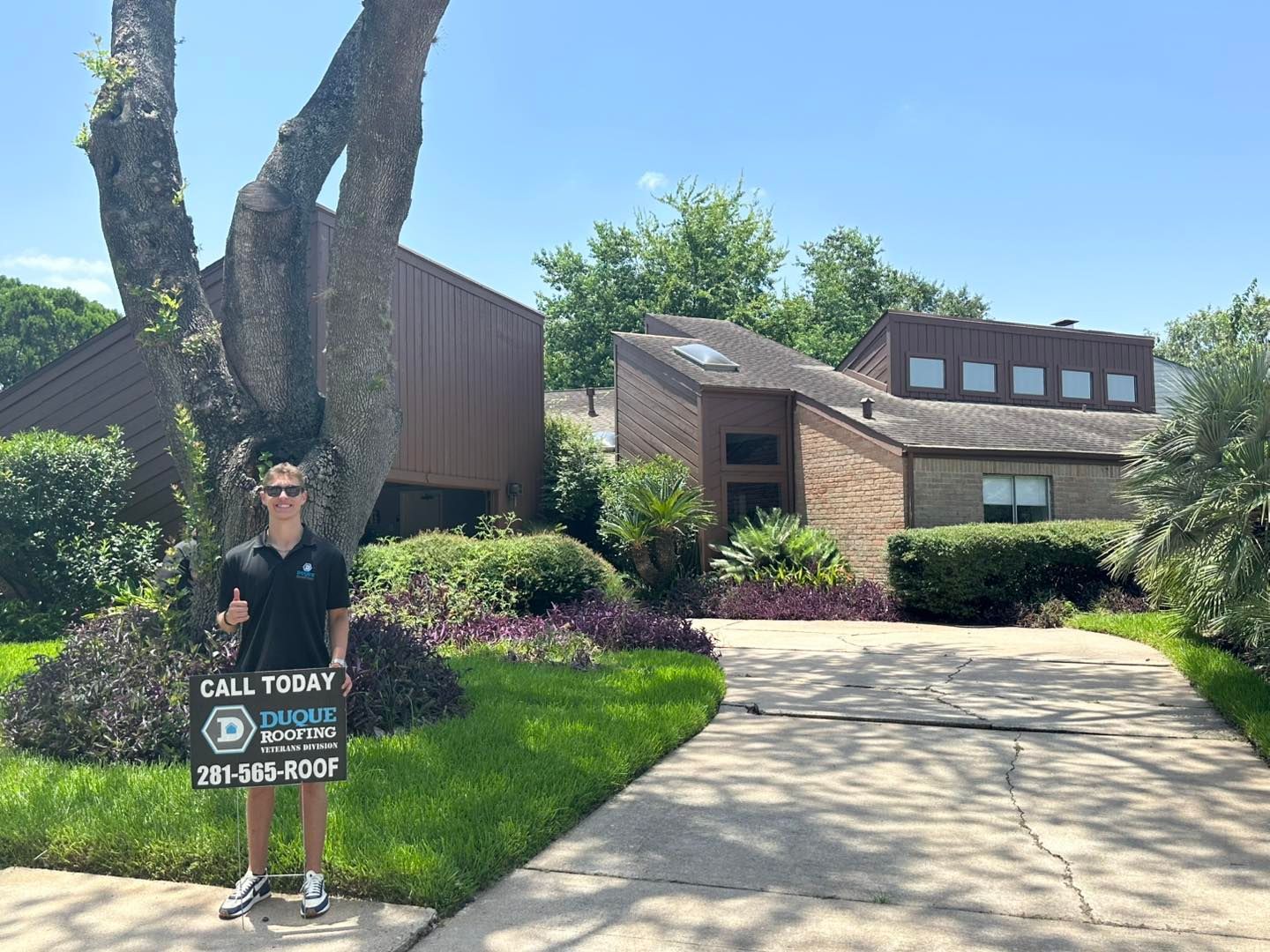 Man holding sign stands in front of a house. Sign advertises roofing services. Sunny day.