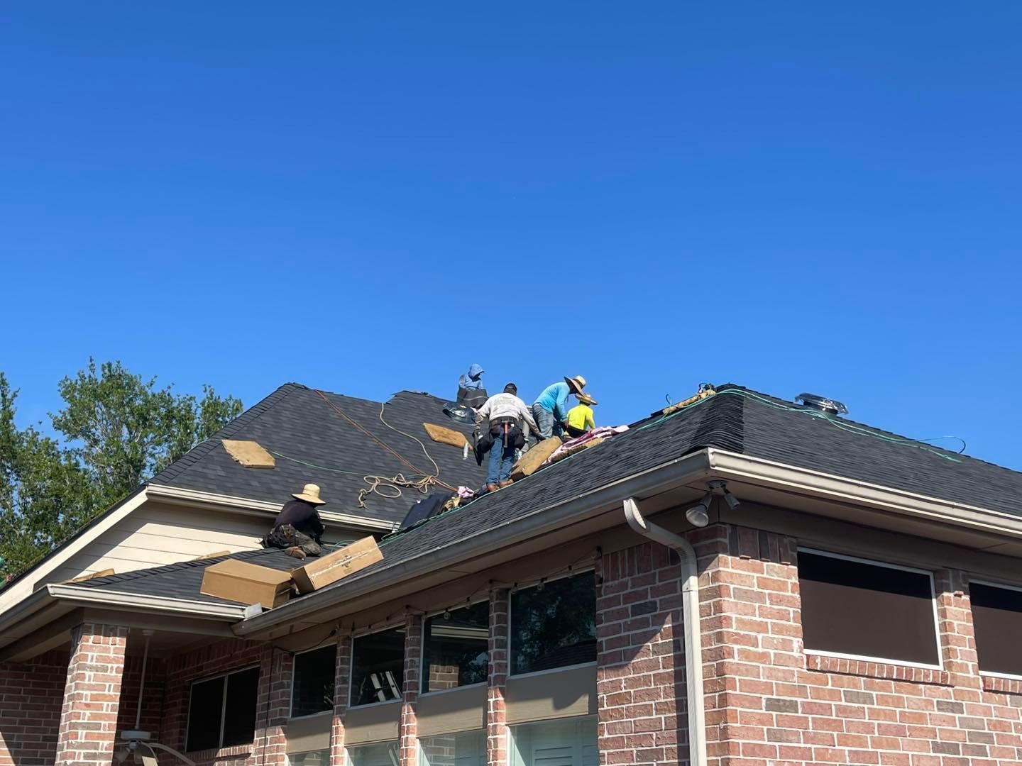 Roofers working on a dark shingled roof under a clear blue sky. Brick home visible.