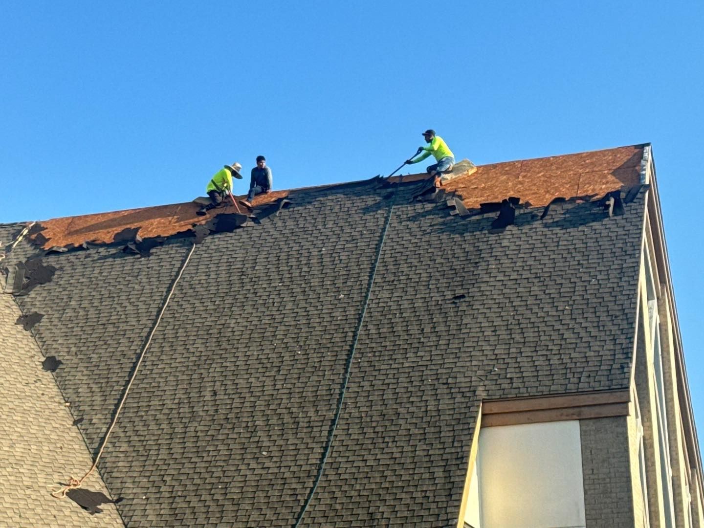Three roofers in yellow vests remove old shingles from a building roof under a blue sky.