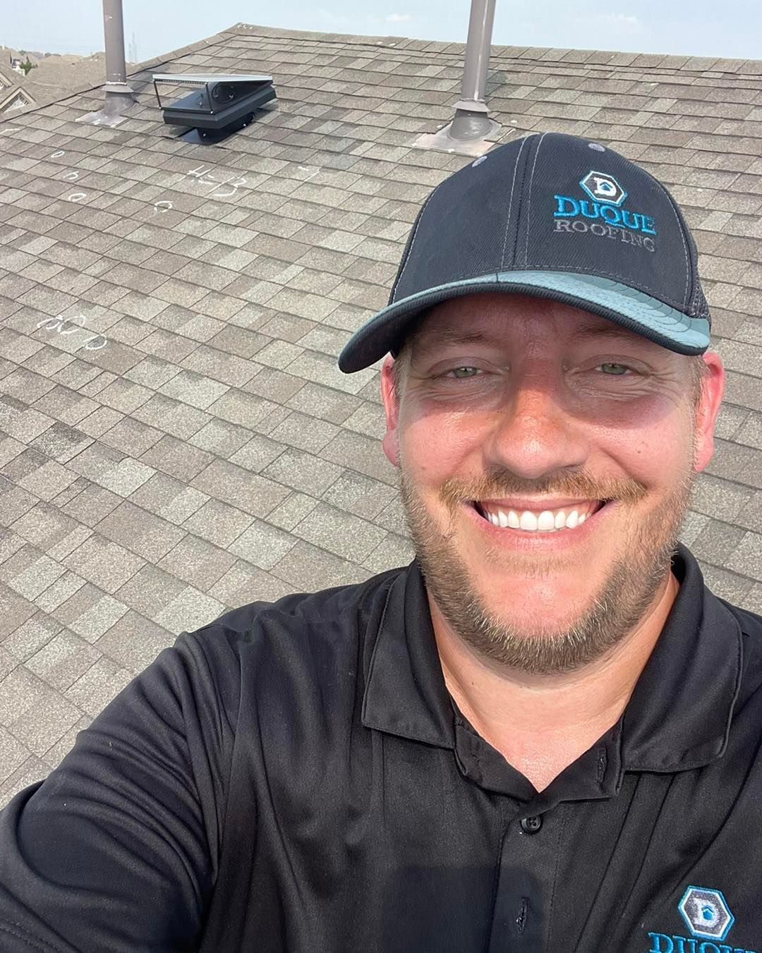 Man in black cap and shirt on a shingled roof, smiling at the camera.
