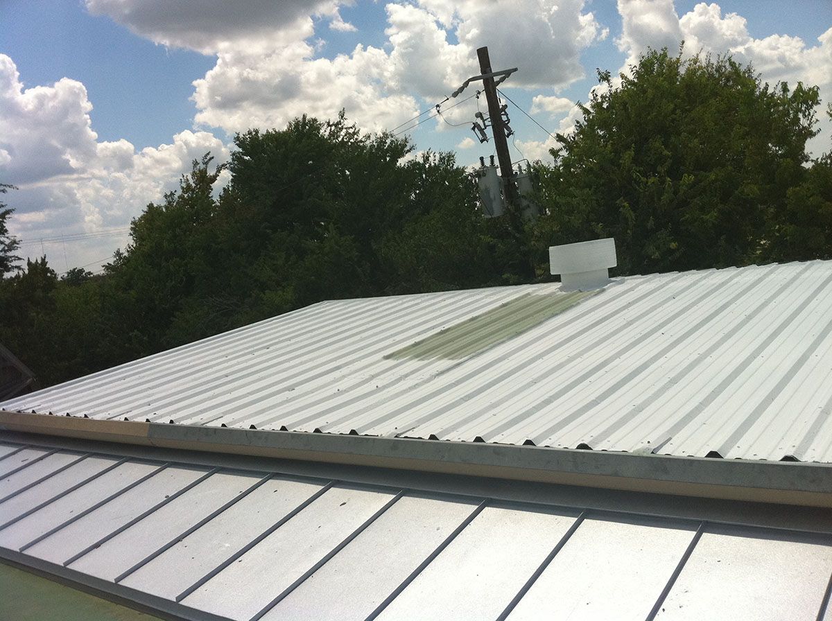 White metal roof with a green patch, vent, and trees against a blue sky with clouds.