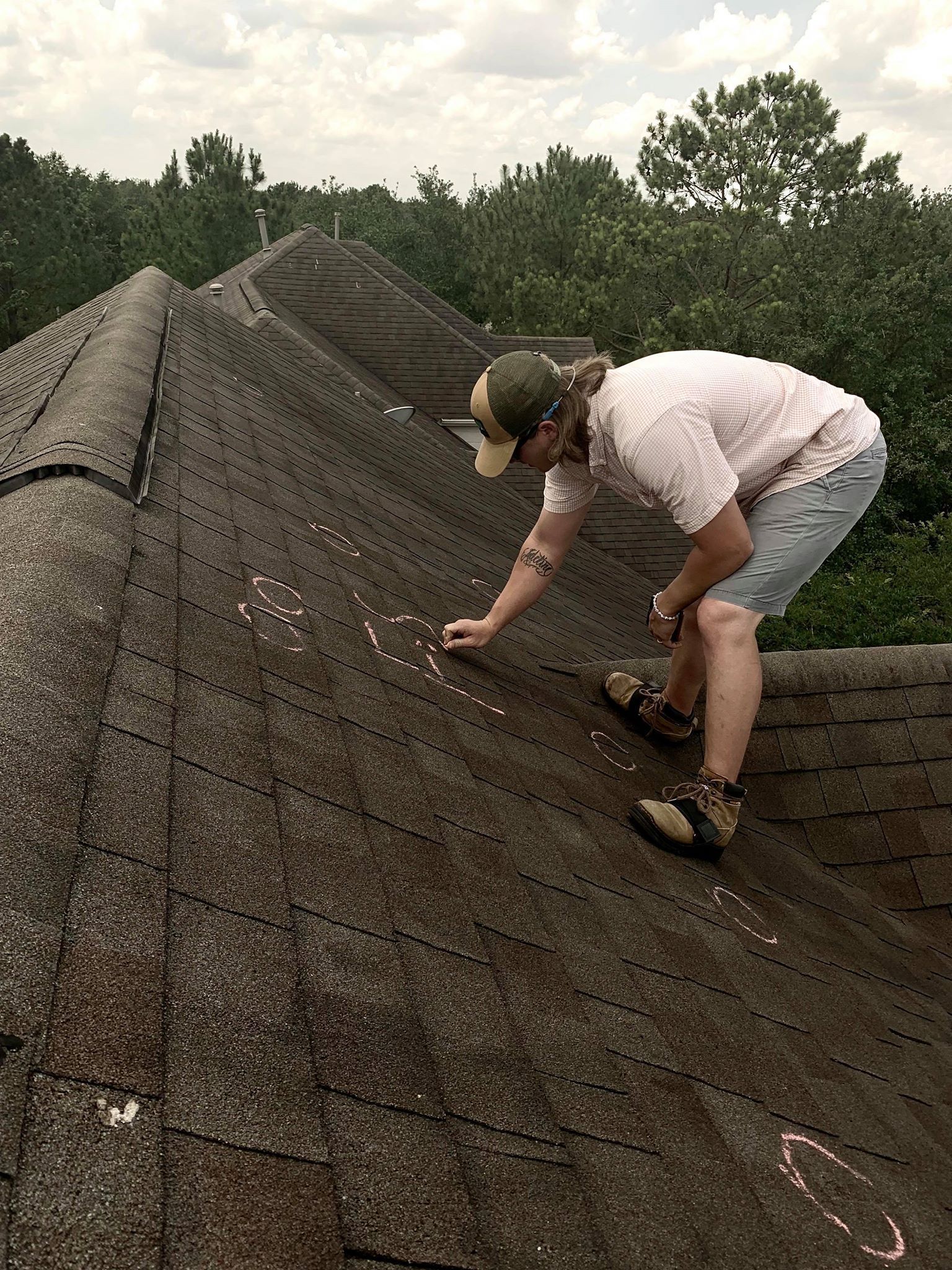 Person on a roof inspecting shingles, marking spots. Overcast sky, trees in the background.