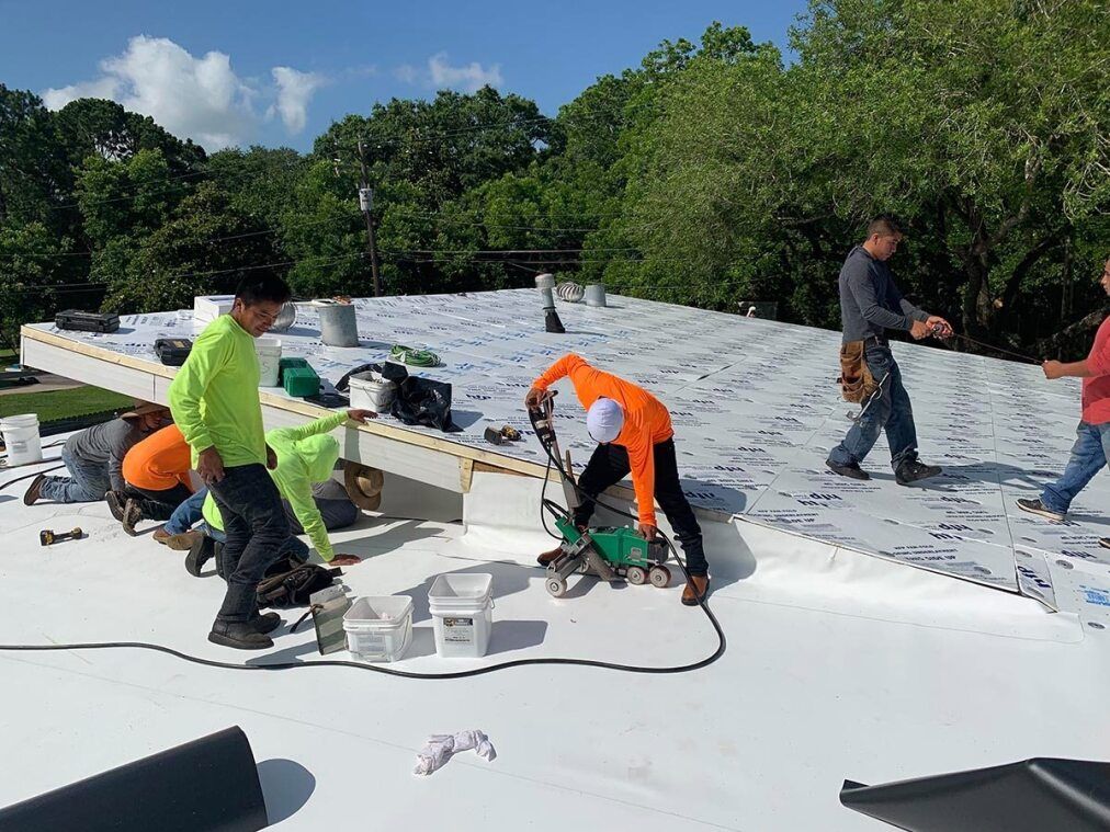 Gray asphalt shingle roof with chimneys and a commercial building in the background on a sunny day.