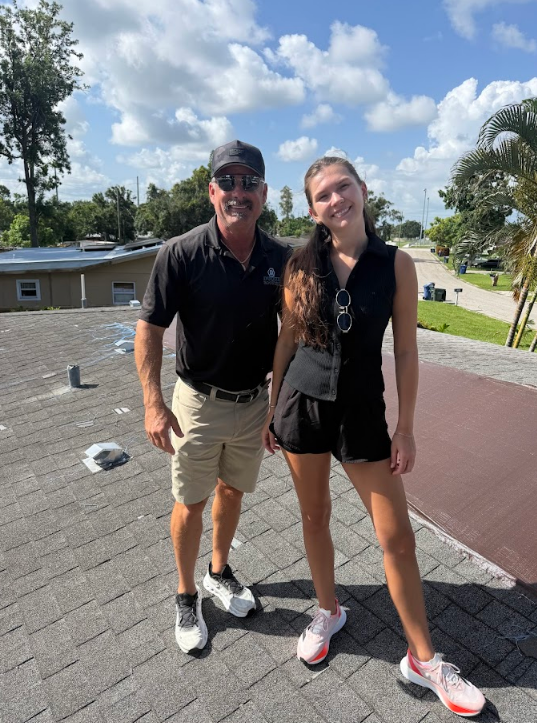 Man in baseball cap smiles on a rooftop; gray shingles and vents visible.