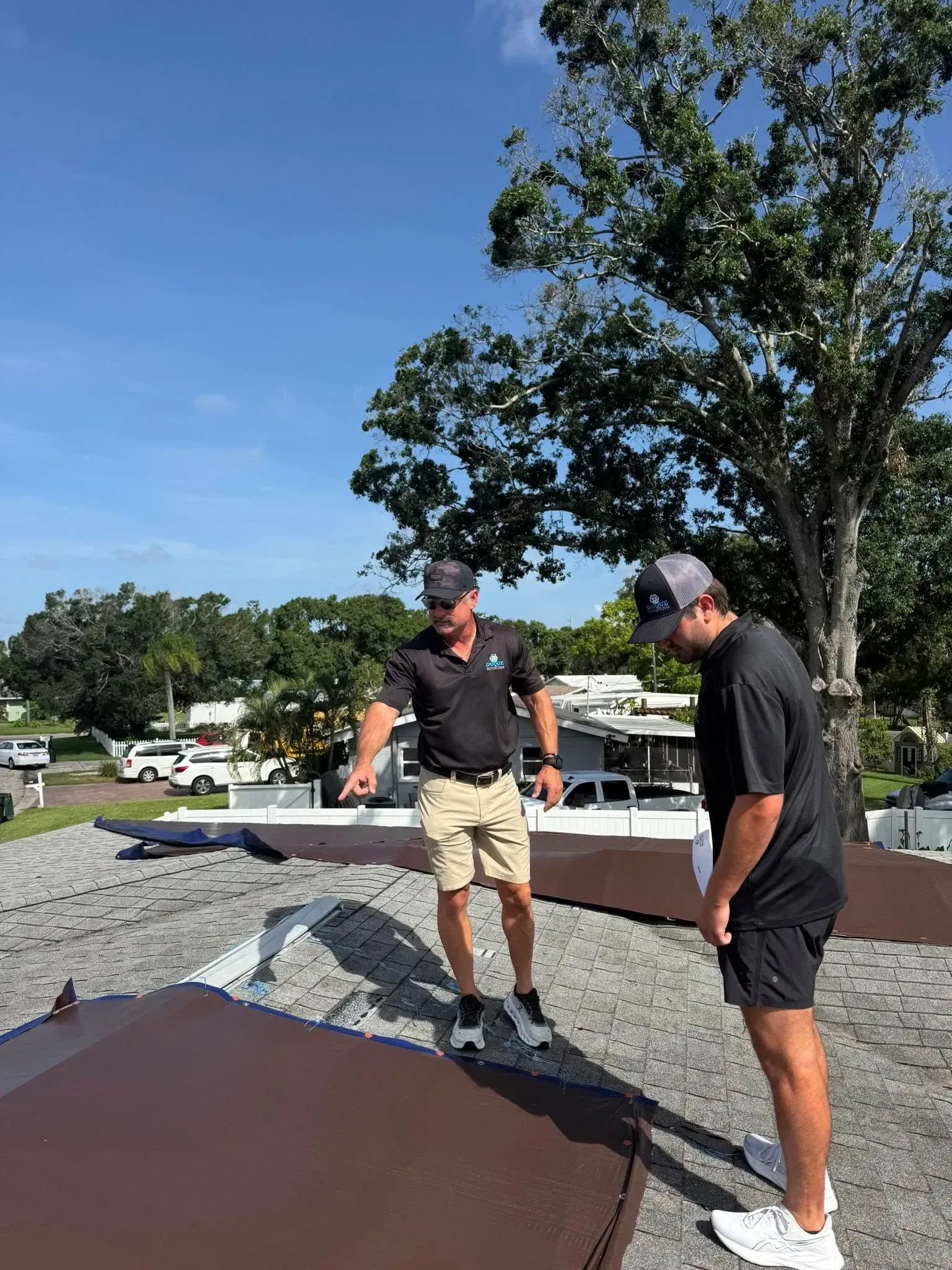 Two roofers installing shingles on a roof under a blue sky with clouds.