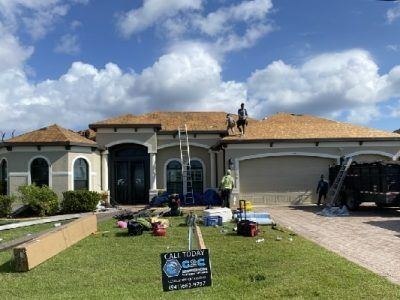 Roofers working on a house with a tan roof and stucco siding under a cloudy blue sky.