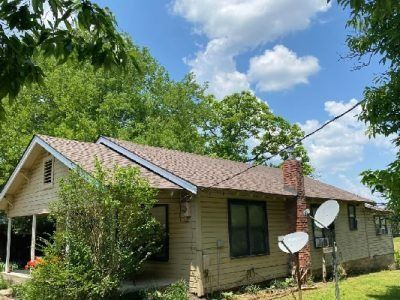 Tan one-story house with brown roof and brick chimney. Two satellite dishes are attached to the side. Green trees and blue sky.