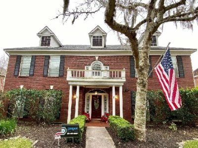 Two-story brick house with shutters, dormers, porch, US flag, tree, and manicured landscaping.
