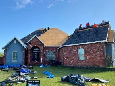 Roofers working on a house with brick and blue siding under a blue sky. Debris is on the lawn.