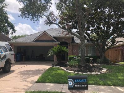 House with dark roof, garage, and landscaping under a tree on a sunny day. A work truck is in the drive.