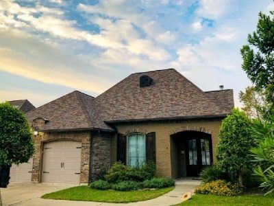 House with brick and beige exterior, brown shingle roof, arched doorway, and garage. Green lawn and shrubs.