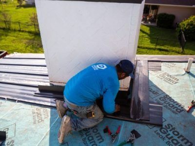 Roofer kneeling, installing flashing around a white chimney on a roof with blue underlayment.