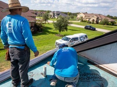 Two roofers in blue shirts working on a roof, with a truck and houses in the background.