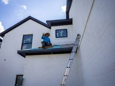 Construction worker on a roof, giving a peace sign. White building with black trim, ladder leaned against the wall.
