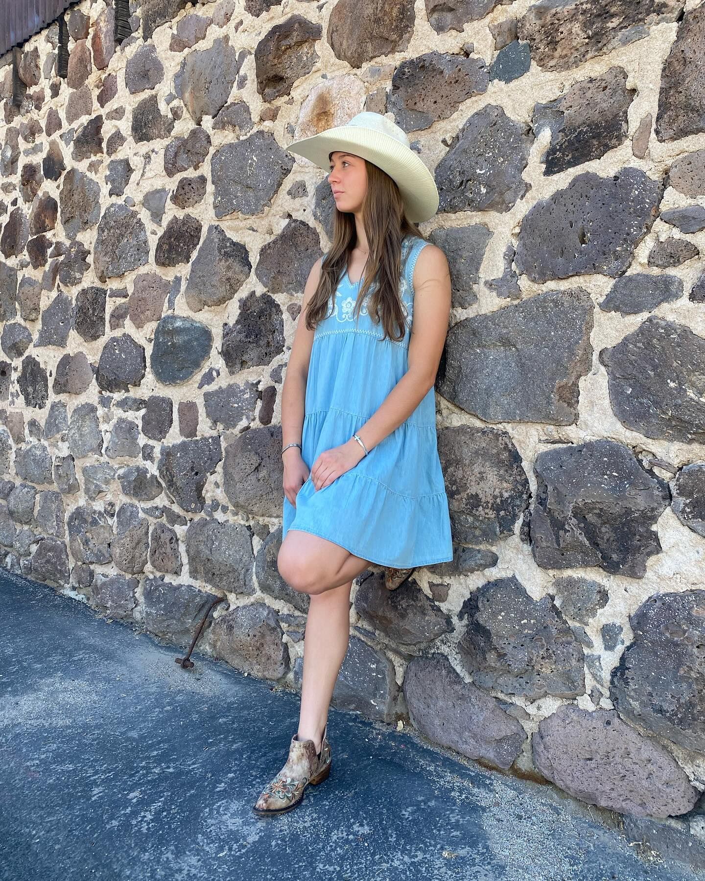 A woman in a blue dress and cowboy hat is leaning against a stone wall.