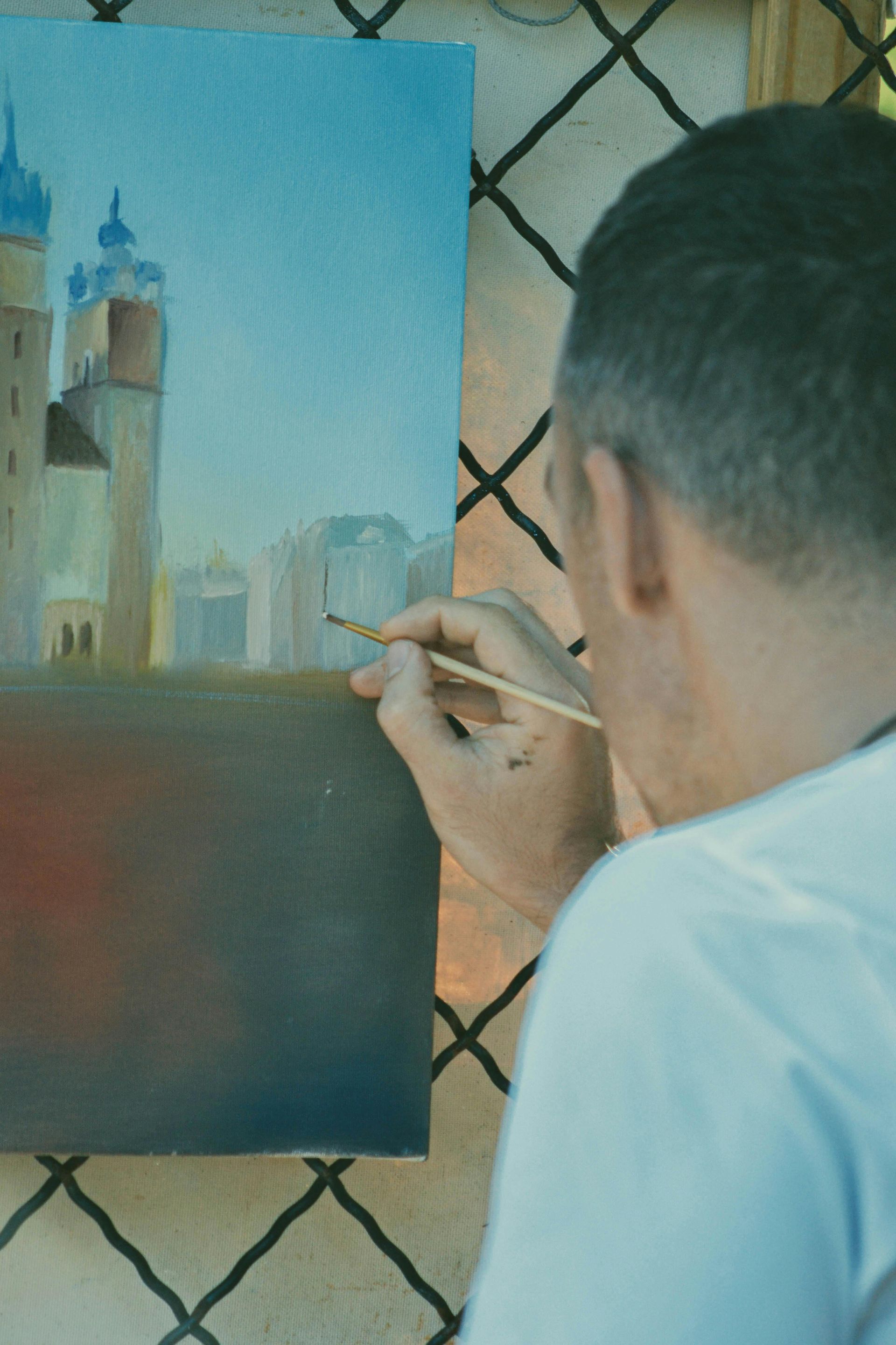 Een kunstenaar schildert een stadsgezicht op een doek, met een kleine toren en een blauwe lucht.