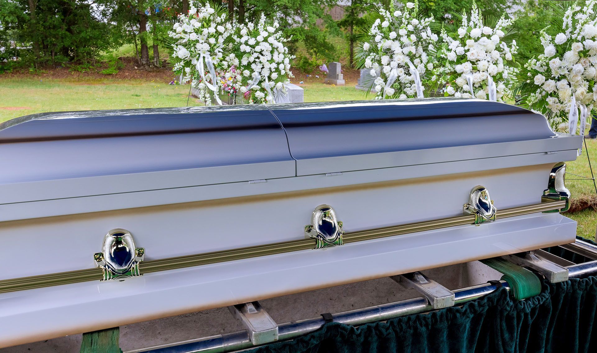 A white coffin is sitting in a cemetery with flowers in the background.
