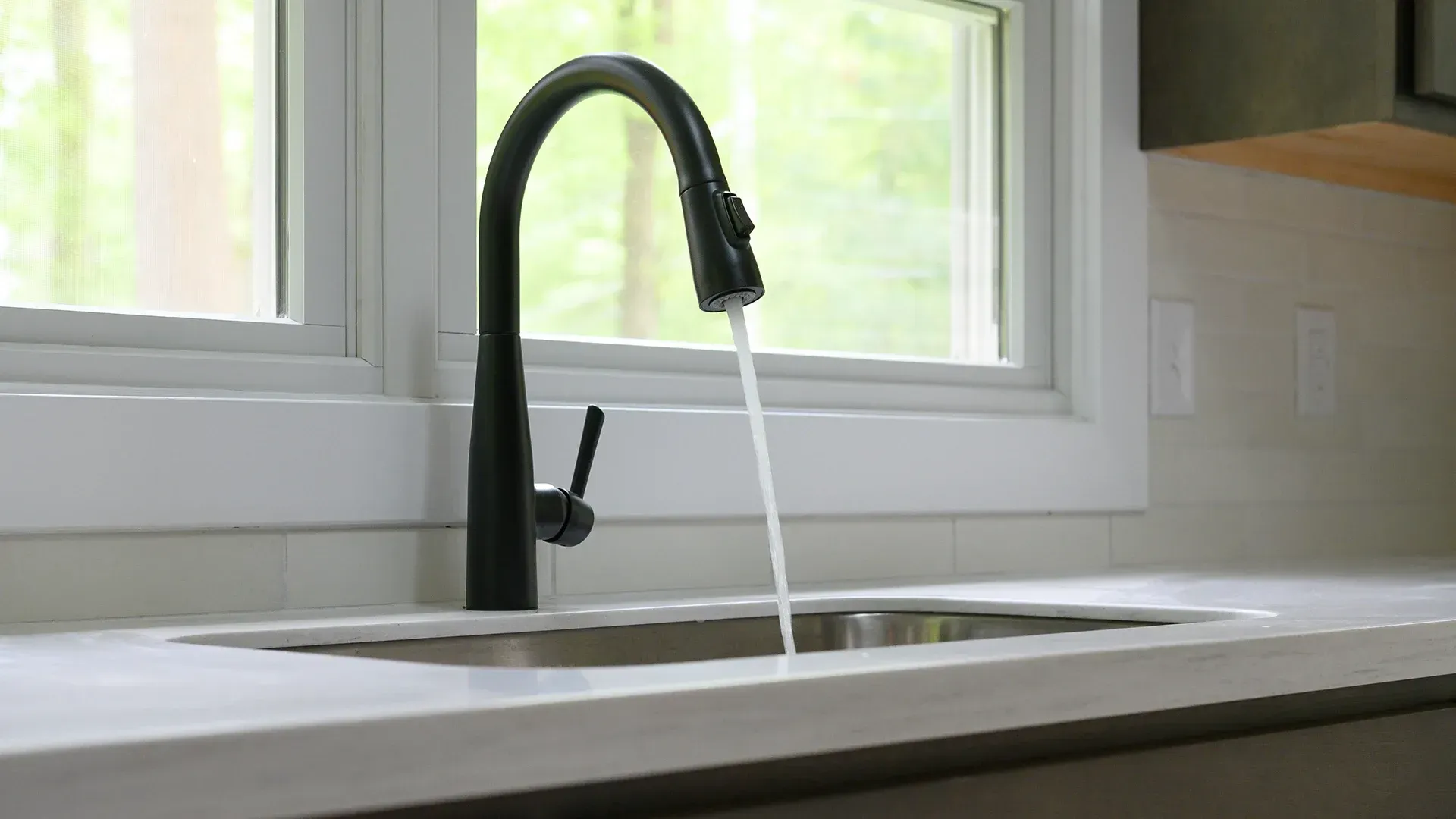 Black kitchen faucet with running water over a stainless steel sink, set in front of a window.