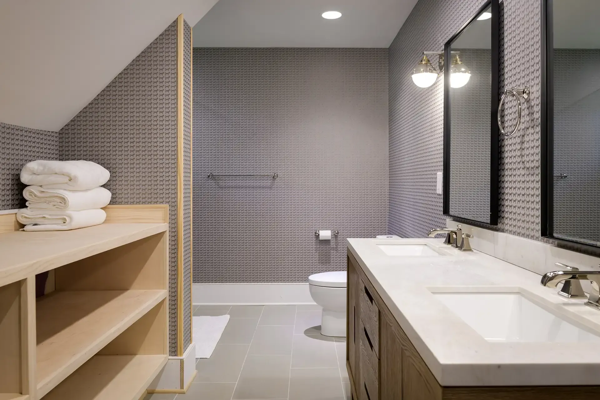 Bathroom with wood vanity, dual sinks, neutral textured wallpaper, and a shelf for towels.