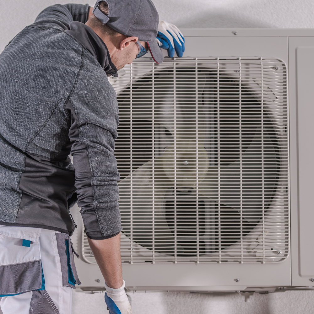 HVAC technician inspects an air conditioning unit outside. White and gray clothes, white gloves, and a blue cap.