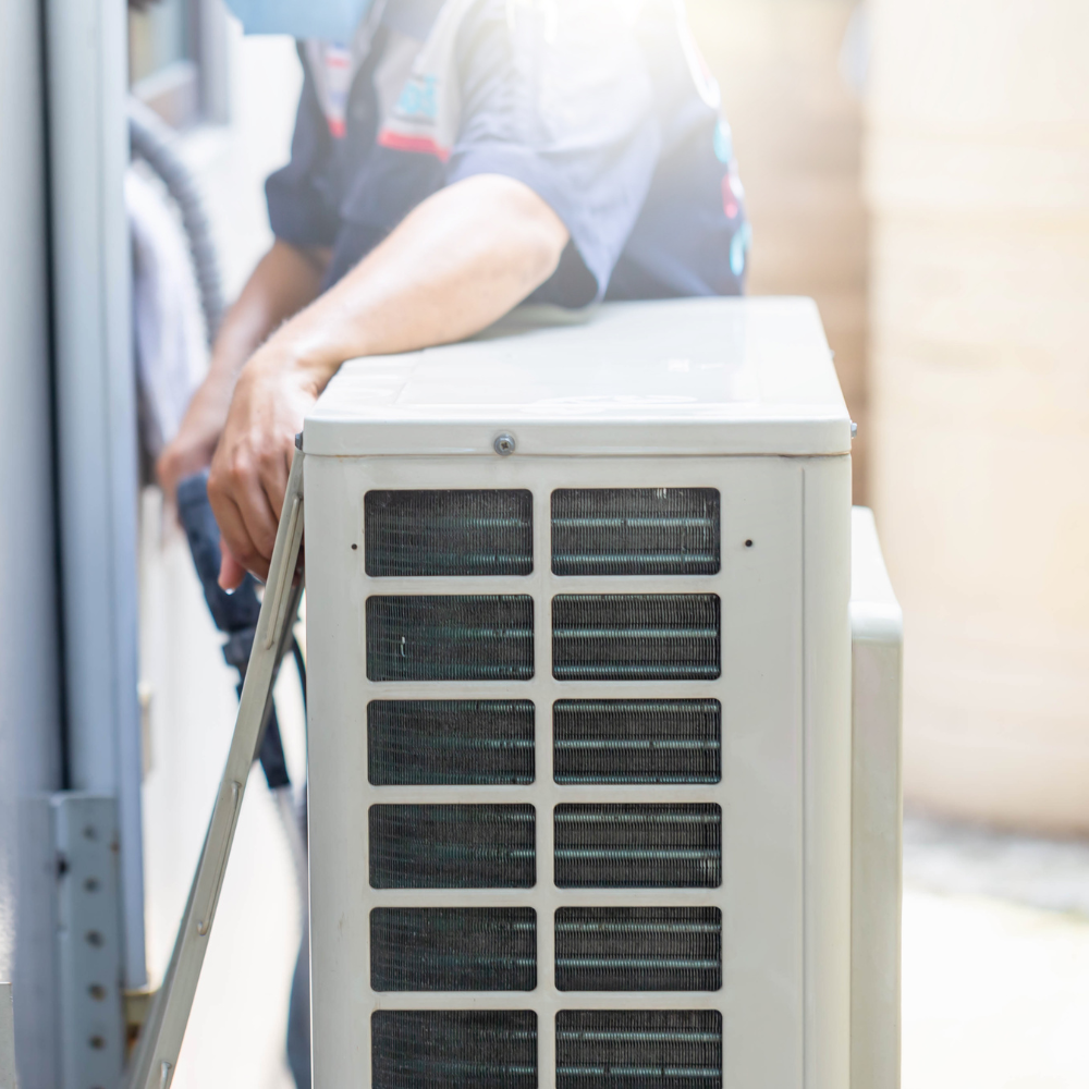 HVAC technician installing an air conditioning unit, outdoors. The unit is white; worker wears a mask.
