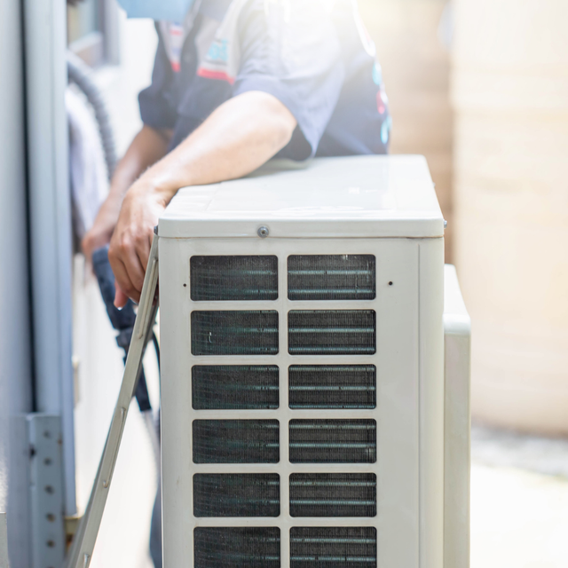 HVAC technician installing an air conditioning unit, outdoors. The unit is white; worker wears a mask.