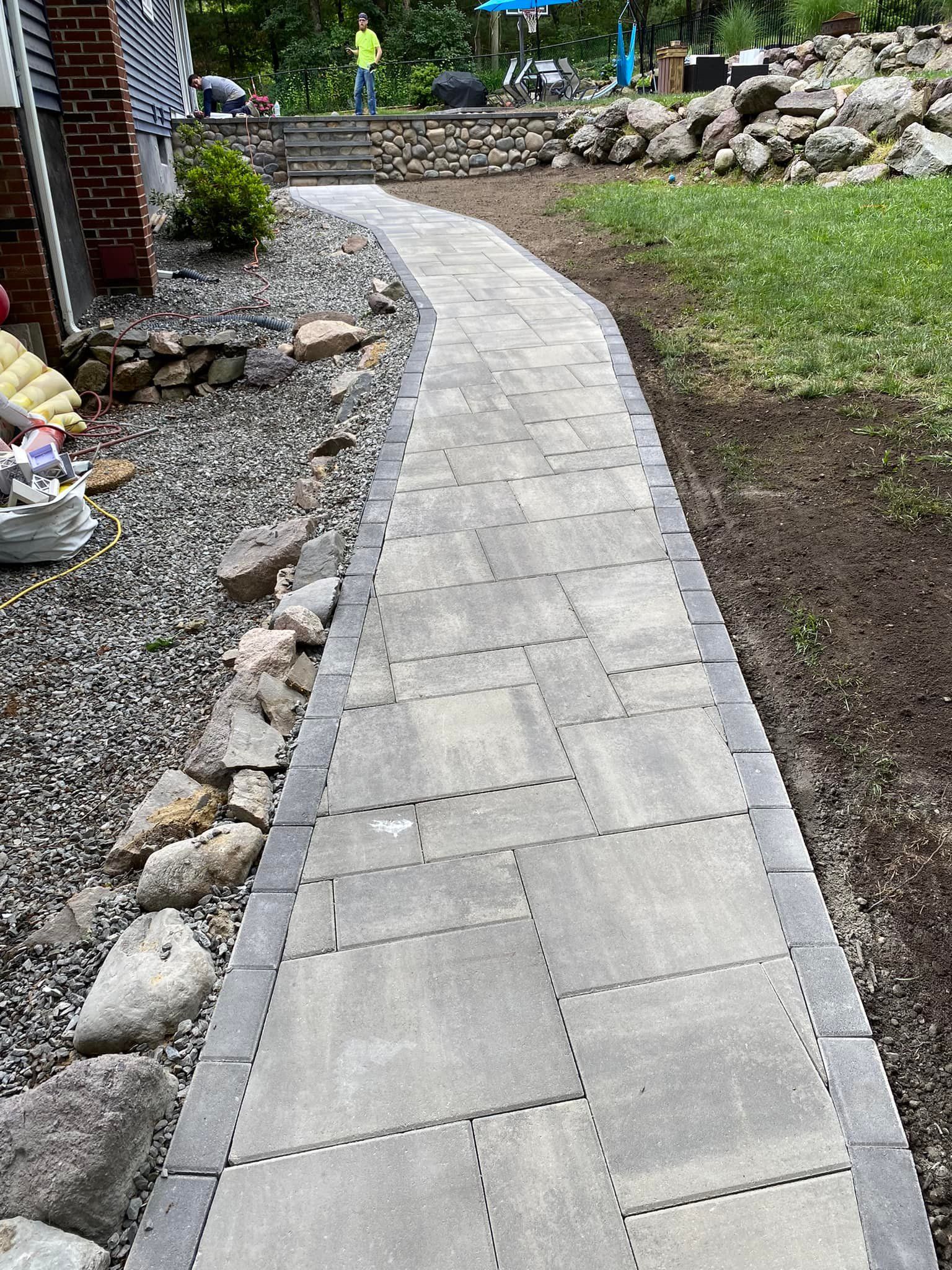 A stone walkway curves through a yard. Gray pavers are bordered by darker stones and gravel.