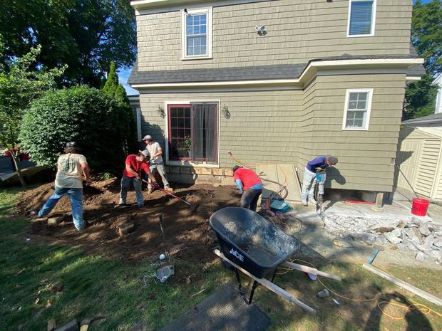 Construction workers laying patio stones next to a house with green siding; wheelbarrow in foreground.
