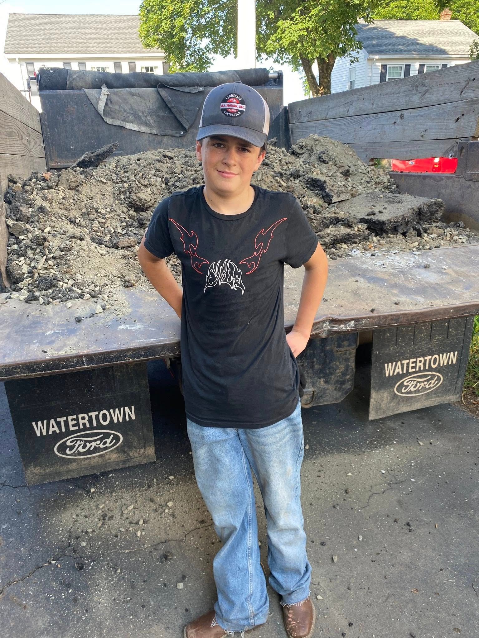 Boy in a baseball cap stands by a dump truck filled with asphalt, hands in pockets.