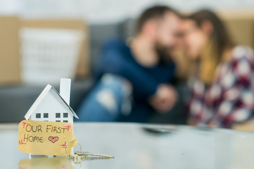 A couple is sitting on a couch with a model house and keys on a table.