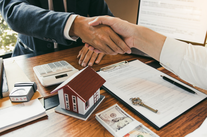 A man and a woman are shaking hands over a table with a model house and money.